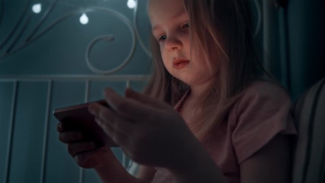 Little girl is looking at the phone. The child is playing on the smartphone. A girl uses the phone in the dark before going to bed. 4k