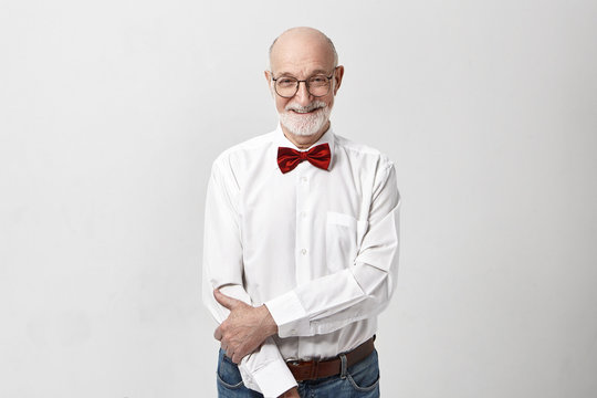 Horizontal image of handsome cheerful Caucasian elderly mature male with bald head and thick gray beard posing in studio wearing red bow tie, eyeglasses and white shirt having cute shy smile