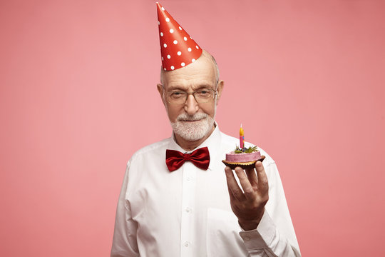 Portrait Of Fashionable Elegant Mature Sixty Year Old Bearded European Male Wearing Red Bow Tie, Glasses And Holiday Hat Posing At Pinkish Studio Wall Holding Birthday Cake With One Single Candle