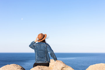 Closeup back view of woman in jeans jacket and hat sitting and looking at blue ocean and sky. - 260843430