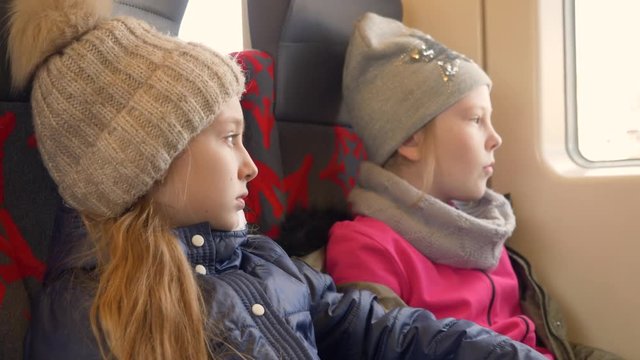 Portrait of two kids girls in winter clothes jackets and caps are travelling by train and looking at window. Side view.
