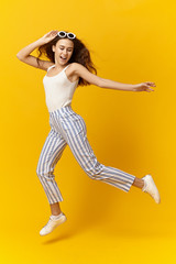 Fototapeta na wymiar Colorful studio picture of trendy fashionable young female with loose hair and slim body enjoying leisure time jumping or dancing at yellow wall, looking down and laughing gaily, having carefree smile
