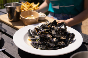 A big bowl of mussels in a creamy sauce served to a woman with fries in the sunshine