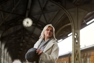 A blonde girl in a white coat stands in the background of the station and holds a hat. Travelling by train.