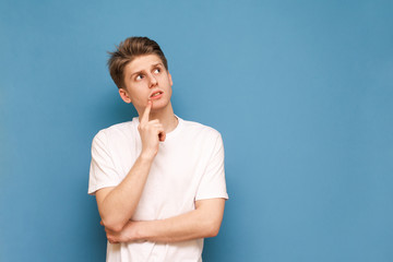 Portrait of a thoughtful young man wears a white T-shirt, holds his finger around his mouth, stands on a blue background and looks at an empty space for advertising. Isolated. Copyspace