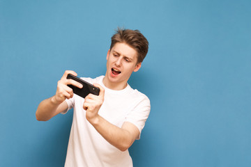 Positive teenage gamer in a white T-shirt plays a game on a smartphone wearing a white T-shirt, isolated on a blue background. Emotional guy playing mobile games. Mobile Gaming Concept