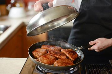 The chef in the kitchen prepares fresh pork steaks on a griddle with steam, the menu is homemade cooking culinary recipes. restaurant menu. Delicious food, recipes and cooking