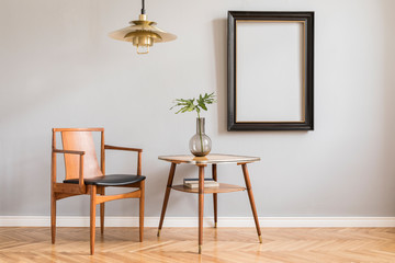 Stylish and bright retro interior with design chair , gold lamp and small table with vase. Black mock up frame on the gray background wall. Minimalistic concept of sitting room. Real photo. 