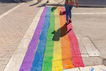 Pride colors paint covers the crosswalk. A woman crosses on the Gay Pride path of the intersection.