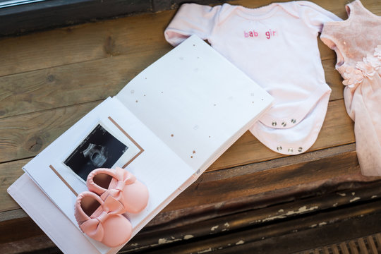 Baby girl pink dress clothes with newborn book album, body and little shoes and ultrasound scanning fetal photo on window wood sill background.