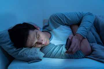 Young caucasian sleepless man bored in bed surfing on the Internet addicted to mobile phone