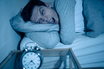 Young man in bed with alarm clock feeling desperate and distress not able to sleep with insomnia