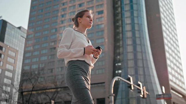 Adult Caucasian Confident Young Business Woman with Hair Bun is Using Smartphone App and Walking Outside by Modern Office Building. Medium Long Low Angle 4K Slow Motion Corporate Tracking 2 in 1 Shot