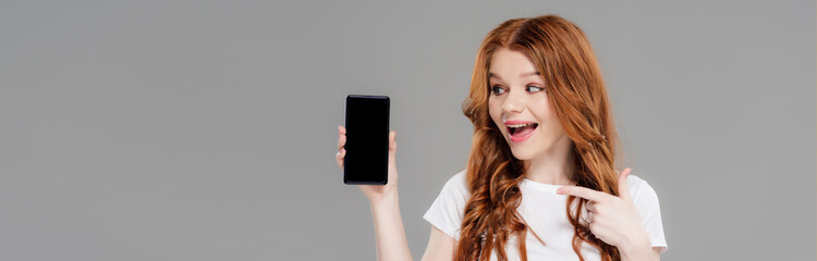 panoramic shot of beautiful excited redhead girl pointing with finger at smartphone with copy space isolated on grey