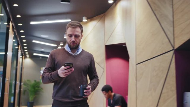 Confident businessman with coffee cup walking along office corridor and text messaging on cell phone. Man passing by espresso machine, meeting colleagues and greeting them