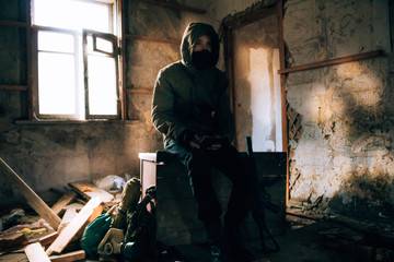 Fototapeta na wymiar Stalker, a soldier guy in uniform and with a gun sits in an old, abandoned house