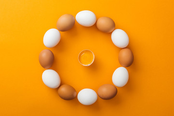 top view of white and brown organic chicken eggs arranged in round frame with smashed one inside on bright orange background