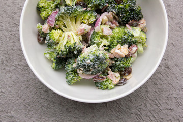Bowl of broccoli crunch salad with red onions, dried cranberries, cashews and bacon against grey...