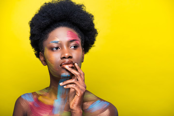 Pensive african american woman with body art touching lips and looking away isolated on yellow