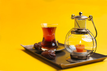 Ramadan Kareem Festive, close up of dates on plate with oriental candle lamp and cup of black tea on yellow background.
