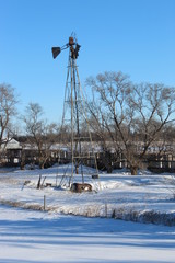 Winter landscape with a old windmill and a old car at its base a couple trees and a old fence.