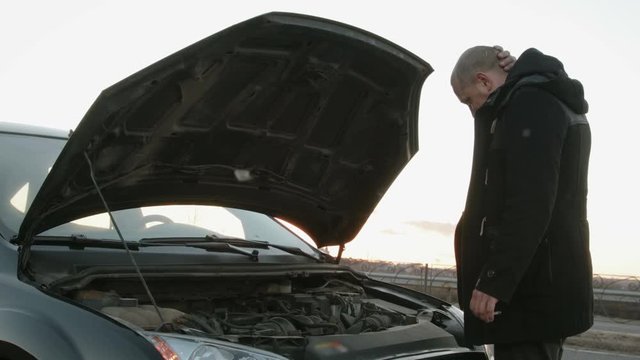 The average plan of a young Caucasian guy with short hair and a Smoking cigarette in his hand, standing near the car black with the hood up. The driver eliminates the damage in the way and scratching