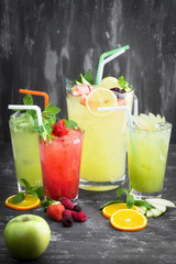 Lemonade in glass pitcher, a glass of lemonade with fresh mint topping,  strawberry frozen and cold iced apple juice. Whole green apple, orange slices and berries on wooden background.