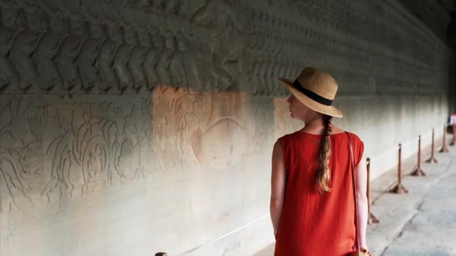Back of the woman in straw hat walking along inner corrdior of Angkor Wat temple and looking at ancient bas-reliefs on the wall. Cambodia