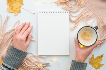 Top view women's hands hold coffe mug and blank notepad on the white table with knitted pink scarf and autumn leaves. Top view. Space for text.