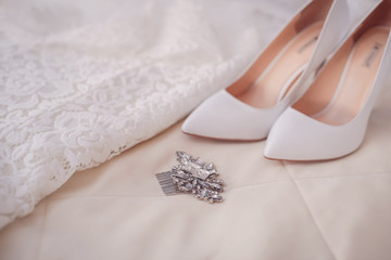 On the beige bedspread lies a wedding lace dress with stylish white sophisticated shoes and a feminine comb hairpin in the hair. Layout morning bride.In warm colors. Womanly and gently.