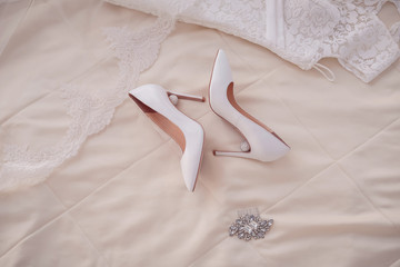On a beige bedspread are beautifully white shoes with heels with crystals, next to a delicate wedding dress and veil with lace and silver hairpin comb with diamonds. Close plan. Gentle morning