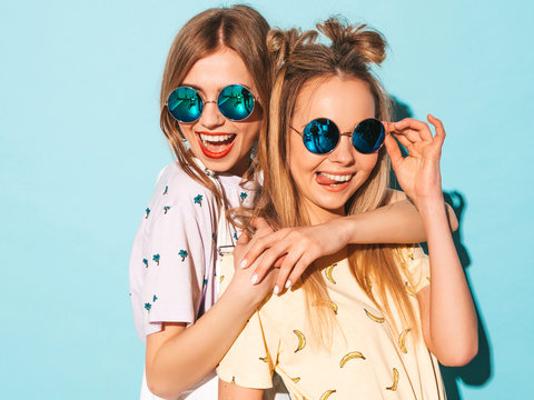 Two young beautiful smiling blond hipster girls in trendy summer jeans skirts clothes. Sexy carefree women posing near blue wall in round sunglasses. Positive models having fun and showing tongue