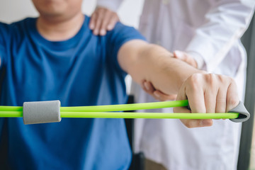 Doctor physiotherapist assisting a male patient while giving exercising treatment on stretching his arm with exercise band in the clinic, Rehabilitation physiotherapy concept