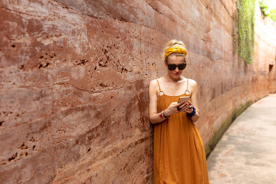 Beautiful blonde woman in summer dress standing by the rammed earth wall and typing on her cell phone.