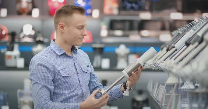 In the appliances store, a man in a shirt chooses a blender to buy by viewing and holding the device.
