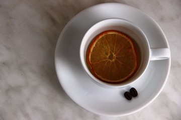 White Cup with slice of dried orange and a couple an coffee beans on a gray table.