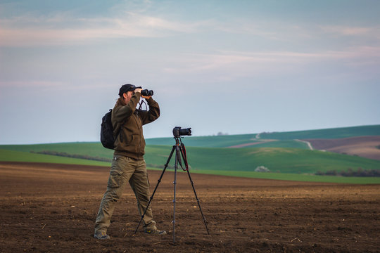 Wildlife photographer looking for animals by binoculars in nature. Rural scene know as Moravian Tuscany at Czech Republic