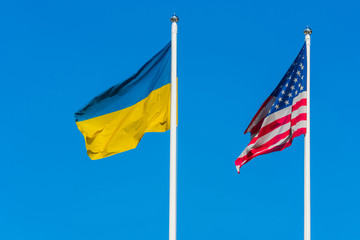 The flag of Ukraine and the flag of the United States of America are sway in the wind next to each other on flagpoles against a background of clear blue sky.