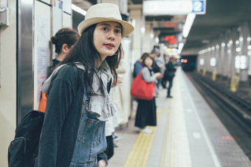 Beautiful young asian woman waiting for train on platform of subway underground station. female backpacker with straw hat looking railway of metro. people commute travel public concept lifestyle.
