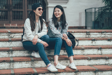 Two cheerful asian girls talking and sitting on stairs in street Santa Barbara County Courthouse. concept of sincere friendship travel together lifestyle. female college students laughing joyful.