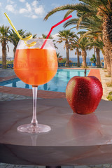 a glass of cold drink and a red apple on the table of the beach cafe against the background of the pool, palm trees and the sea