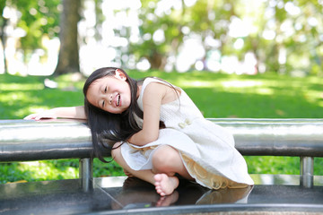 Adorable little Asian kid girl sitting on bench in sunny green park.