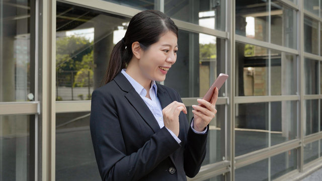 Authentic Lifestyle. Smiling Business Woman Boss Portrait In Black Suit Standing Outside Office Building Skyscraper Texting Message Chatting On Cellphone After Work. Young Lady Worker Enjoy Sunset.