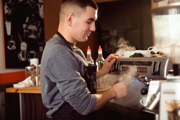 Professional barista holding metal jug warming milk using the coffee machine. Happy young man preparing coffee at counter