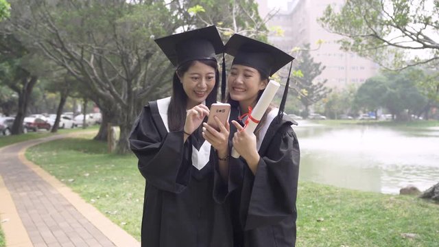 two asian college student girls standing on road in university campus park on sunny day using mobile phone chatting looking screen. young fresh graduates talking about new job interview laughing.