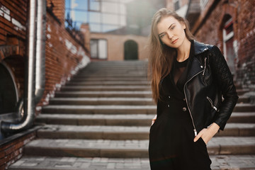 Obraz na płótnie Canvas Young beautiful sexy woman in a black leather jacket, black jeans is standing against the wall of an industrial building of glass and brick on the street.