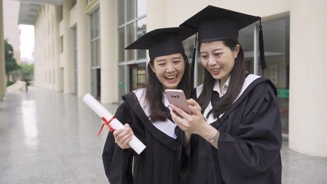 Happy graduation ceremony. Two joyful japanese women in gowns and mortarboard looking at mobile phone screen together and smiling standing out building in campus. asian college student with diploma.