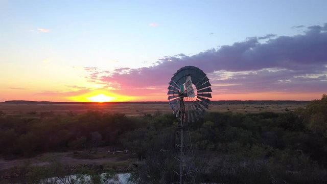 An orbit aerial of a still windmill in the Australian outback, colorful sunset on the horizon.