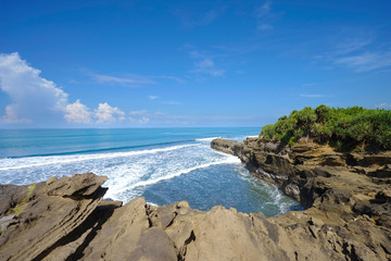 Waves from the ocean to the rock cliffs of the beach on the coast of Bali, with the blue sea and the bright, cloudy sky.