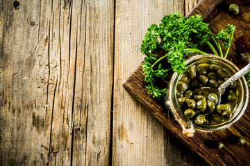 Pickled capers in little jar, with herbs on rustic wooden background copy space